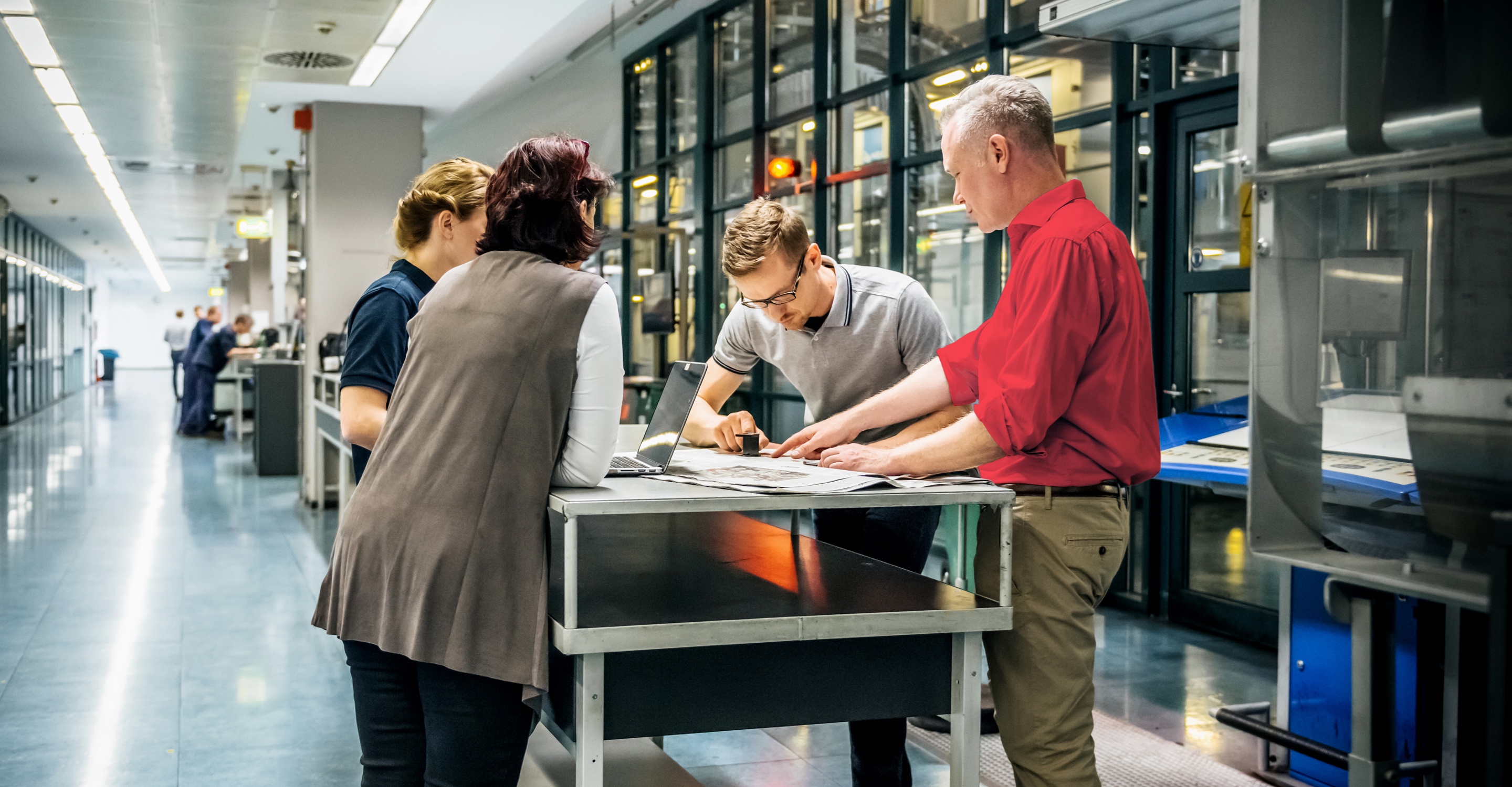 Four engineers surround a work table in what appears to be office space in a factory that can be seen through the windows behind them. One is talking and pointing at papers on the table while the others listen intently