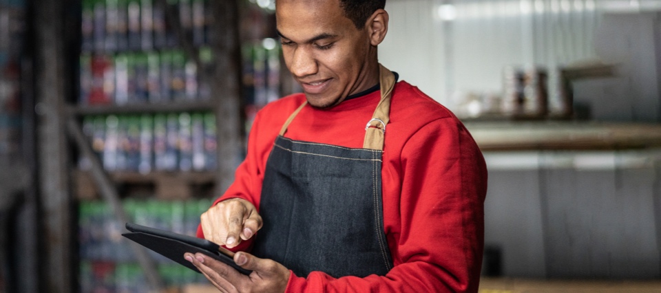 A man in an apron holds a tablet, using his finger to scroll. The background is slightly blurred, but appears to be a warehouse or factory.