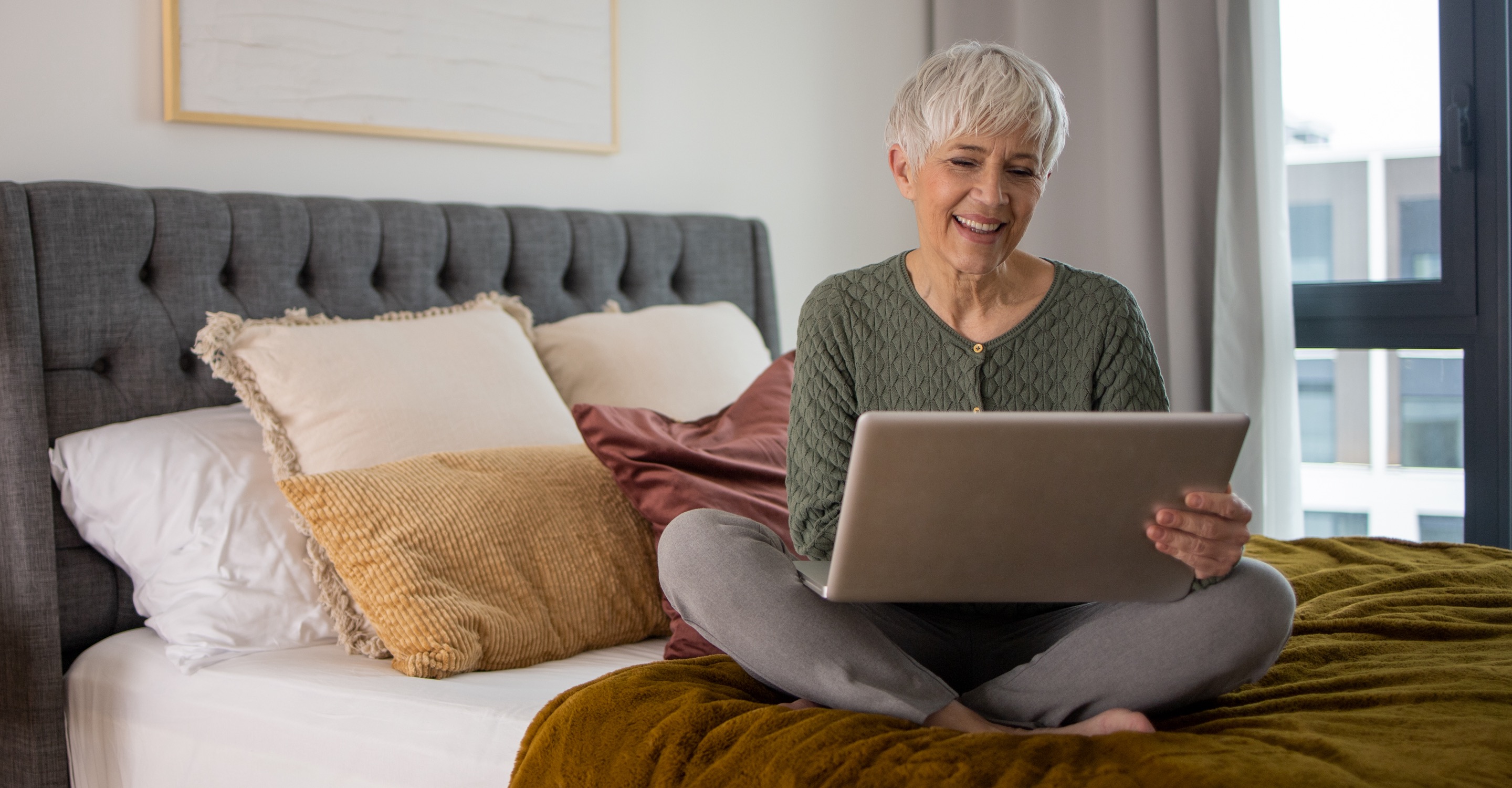 An elderly woman sits cross-legged on a bed, in a brightly lit, modern high-rise apartment bedroom. She smiles as she looks down at the laptop computer open in her lap. 