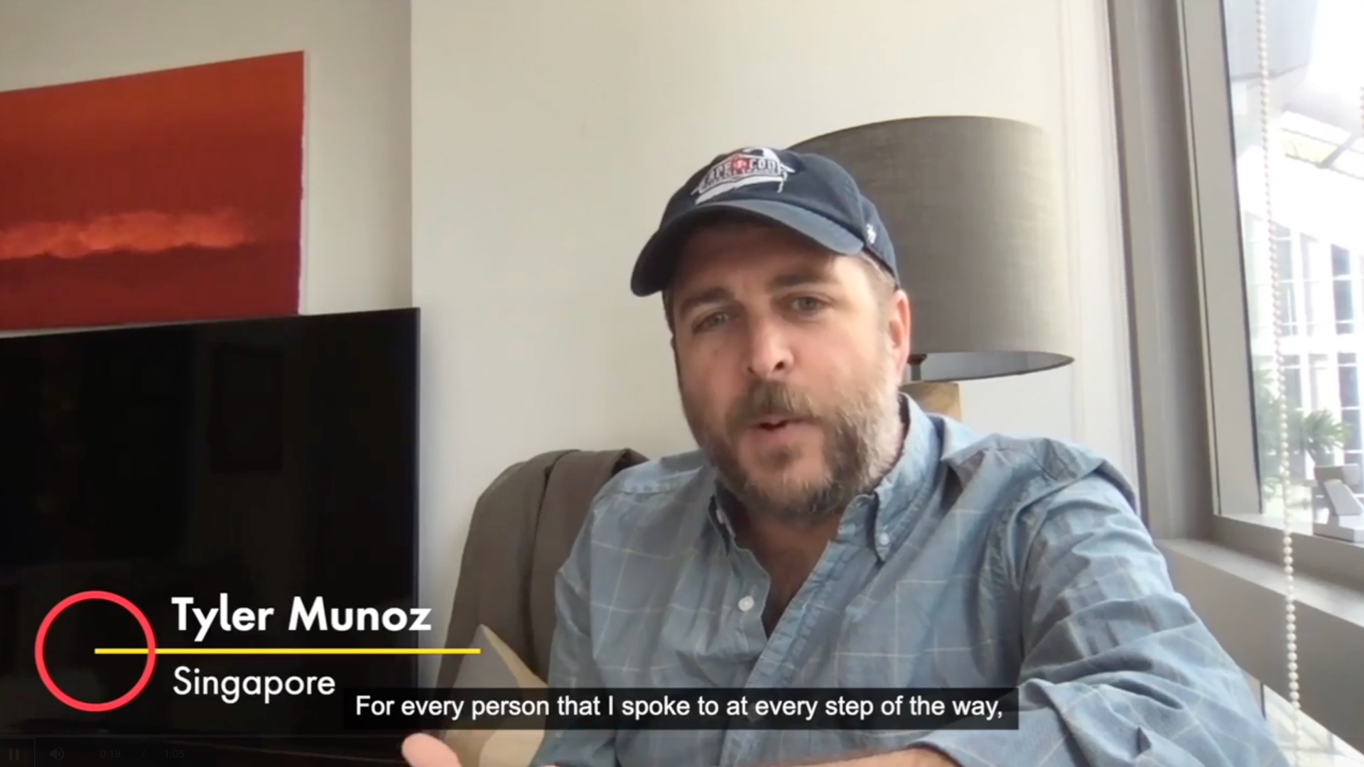 A man in a button up shirt and baseball cap sits in a chair next to a window.