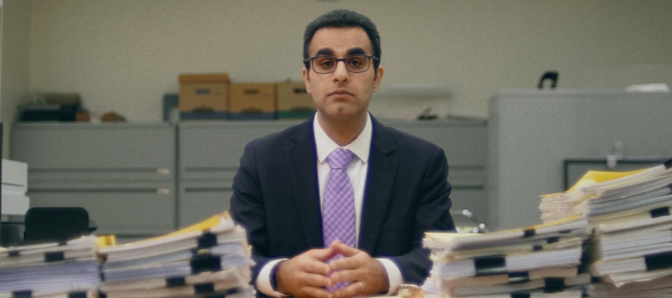 A man with a somber expression wearing a suit sits at a desk surrounded by piles of paper files.