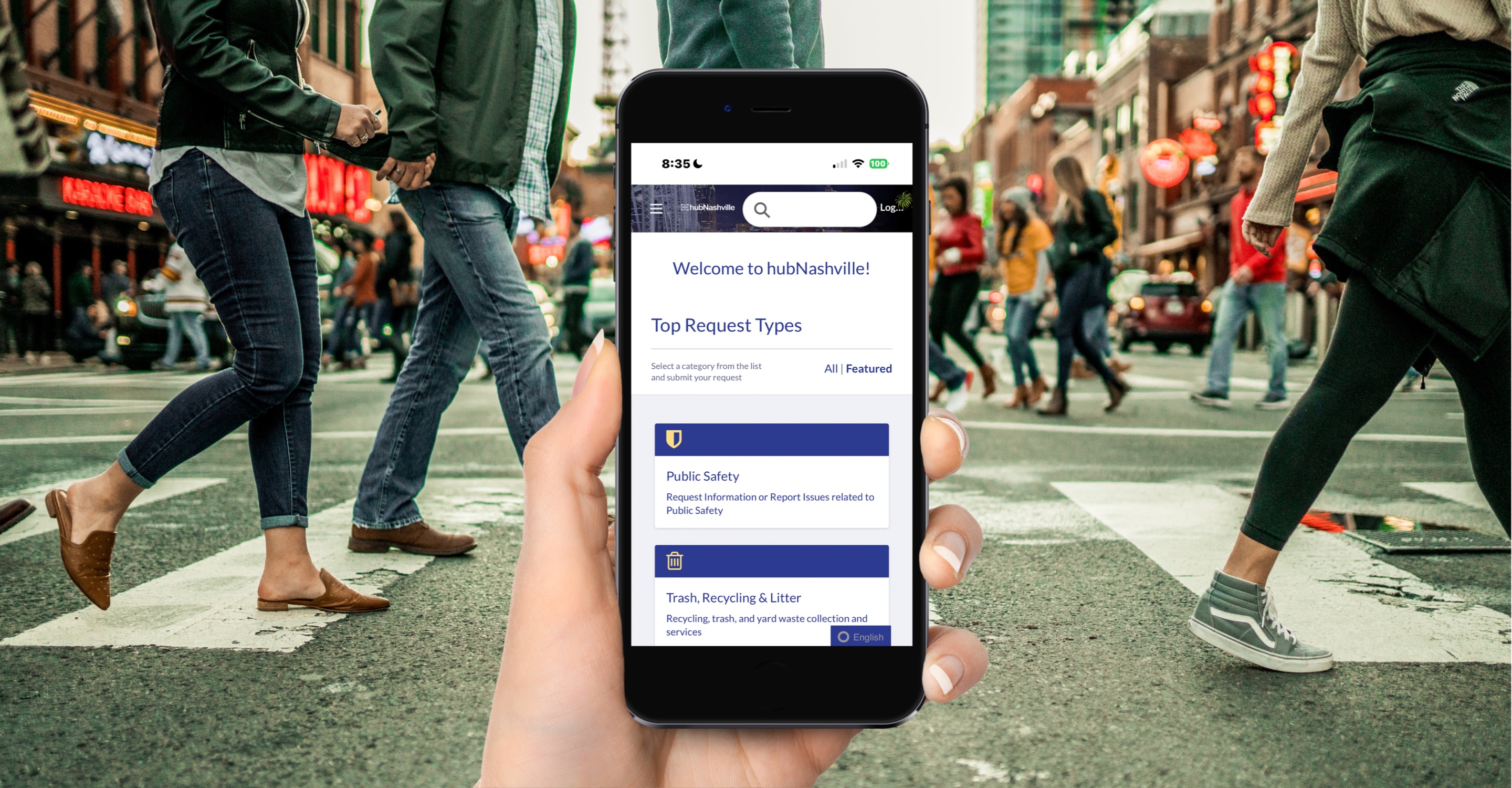 In the foreground, a hand holds up a cell phone close to camera showing the front page of the hubNashville 311 app interface. In the background, people cross the street in a busy, frenetic intersection on Broadway Avenue.