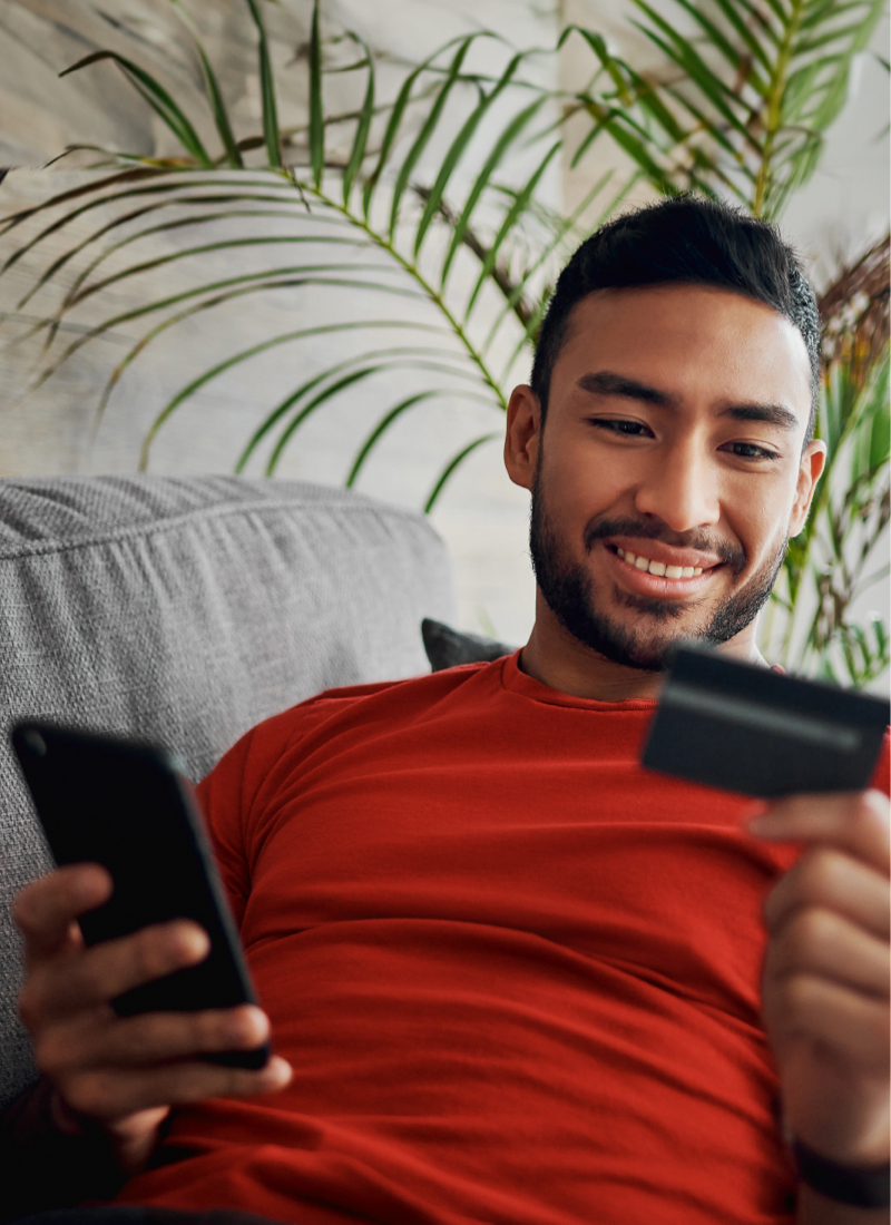 A man sits on the couch holding his cell phone in one hand while looking at his credit card in the other, seemingly making a purchase.