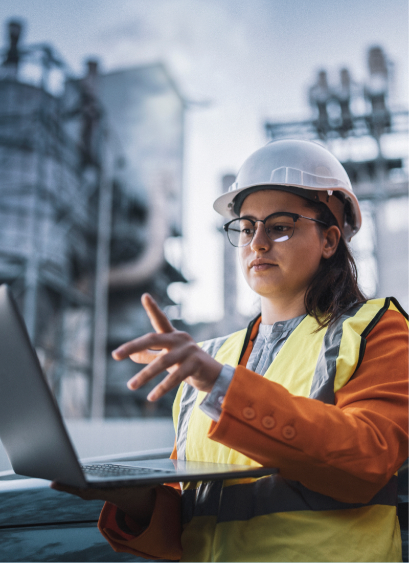 A woman in a high visibility safety vest and helmet holds a laptop in front of a power plant. 