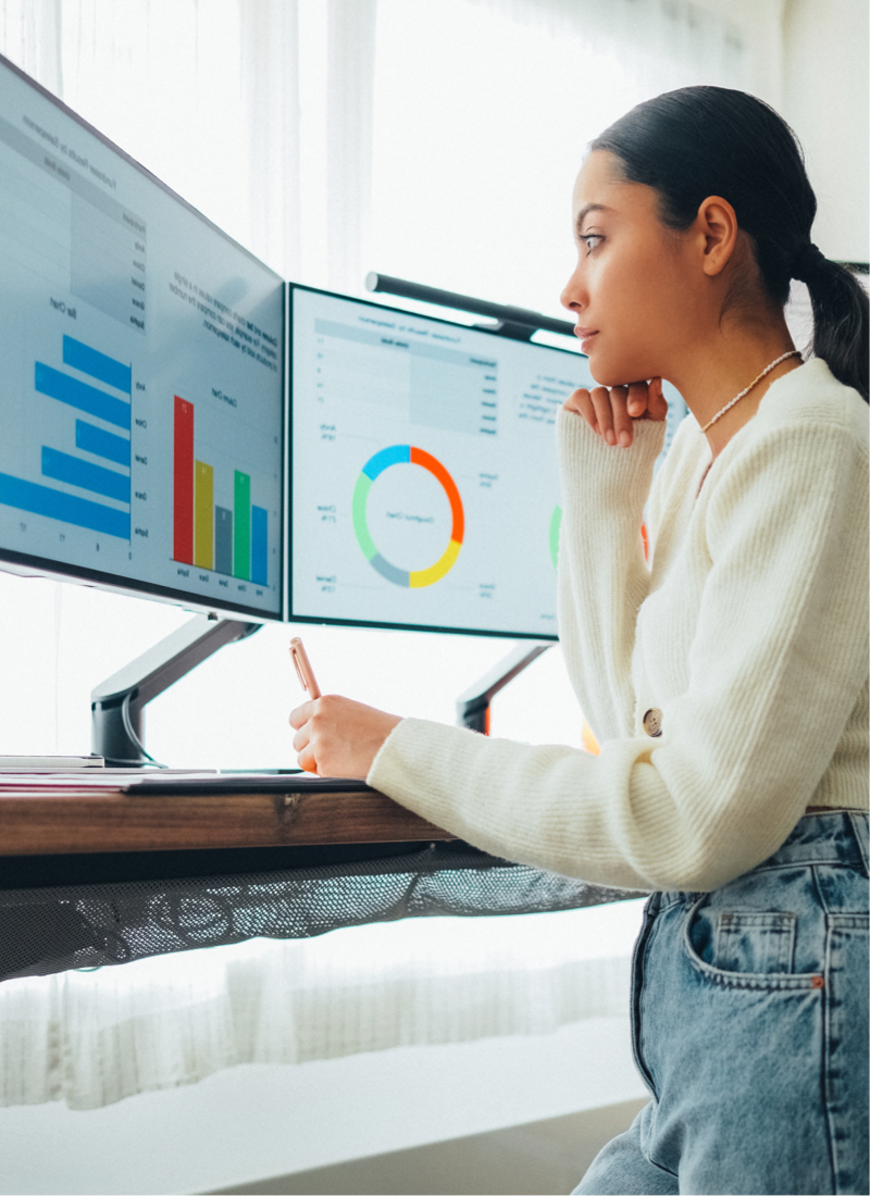 A woman at a standing desk thoughtfully reviews graphs and charts on her desktop computer monitors. 