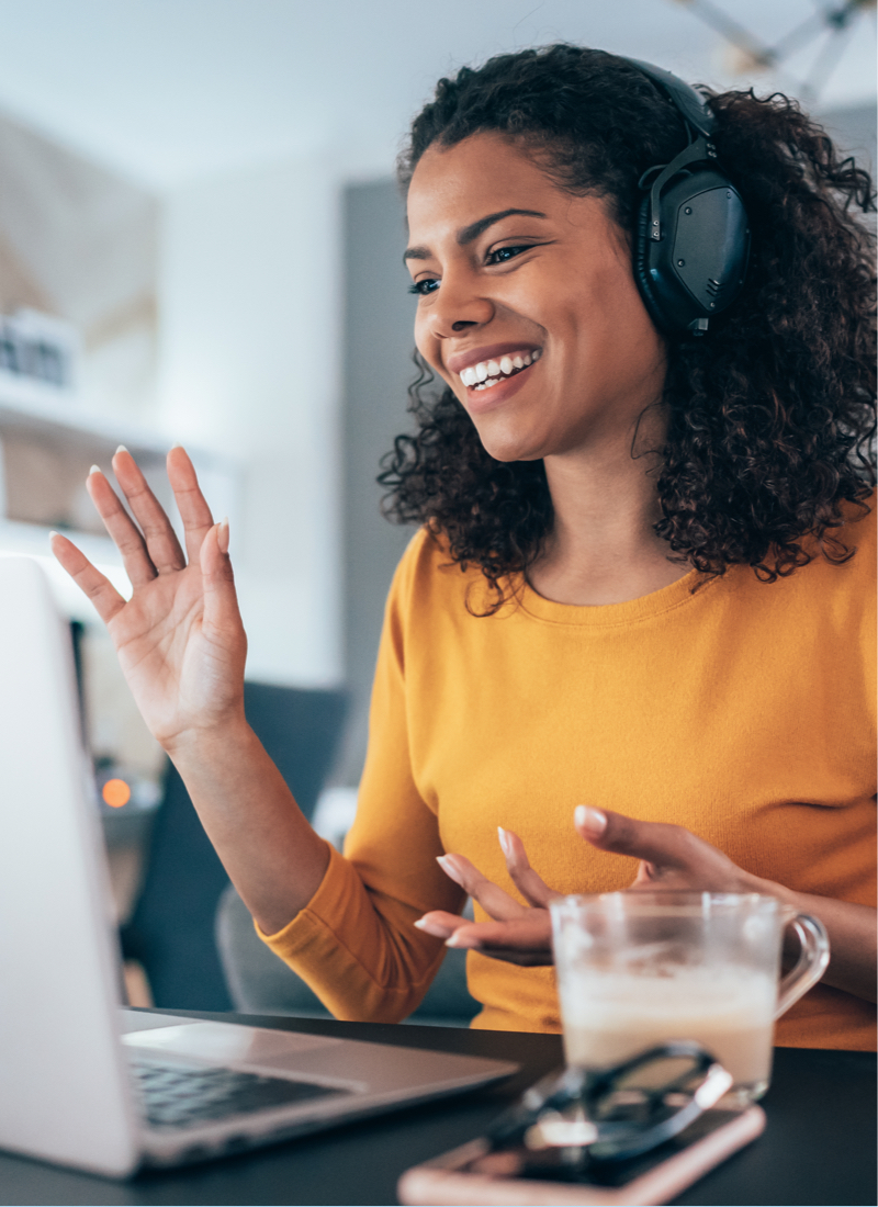 A smiling woman waves at her laptop while wearing over-ear headphones.