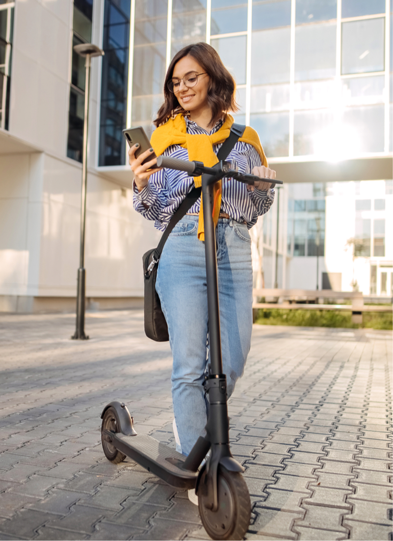 A woman in business casual attire stands on a city street looking at her cell phone in one hand, and holding onto an electric scooter in the other.