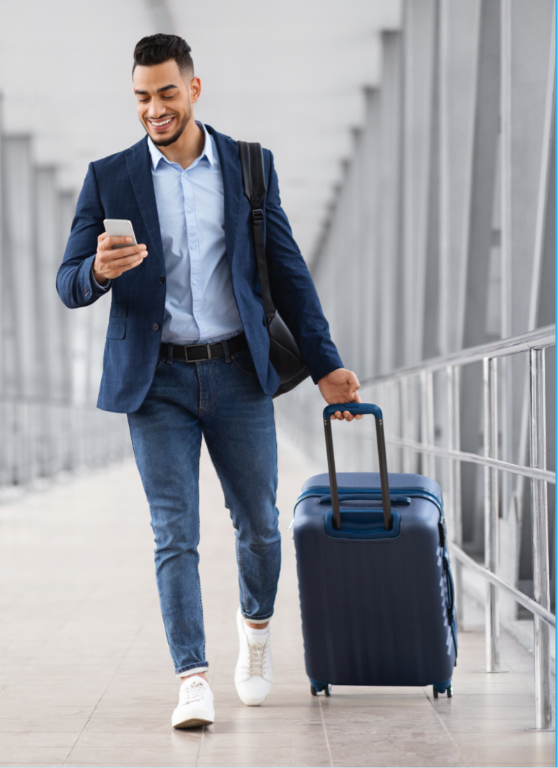 A man in business casual attire walks through an airport hallway, looking at his cell phone in one hand and pulling his carry on suitcase with the other. 